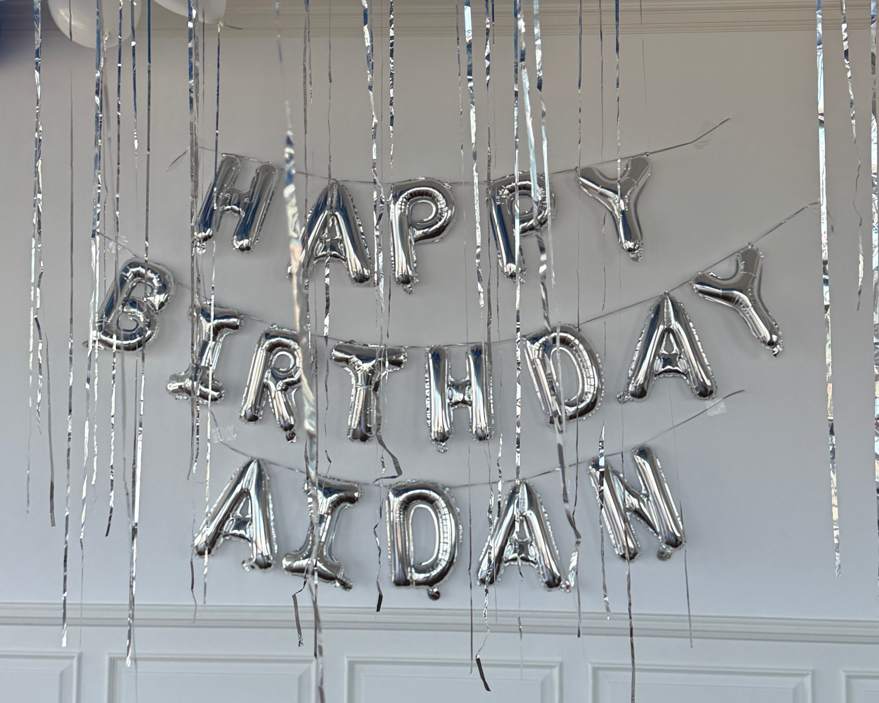 Silver 'Happy Birthday Axdan' balloon banner against a white wall with decorative string.